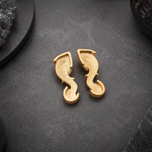 May include: A pair of gold-toned earrings featuring a curved, elongated shape with a textured, matte finish. The earrings have a hollow, recessed center design that tapers into a rounded, hook-like bottom section, presenting a distinct metallic luster.