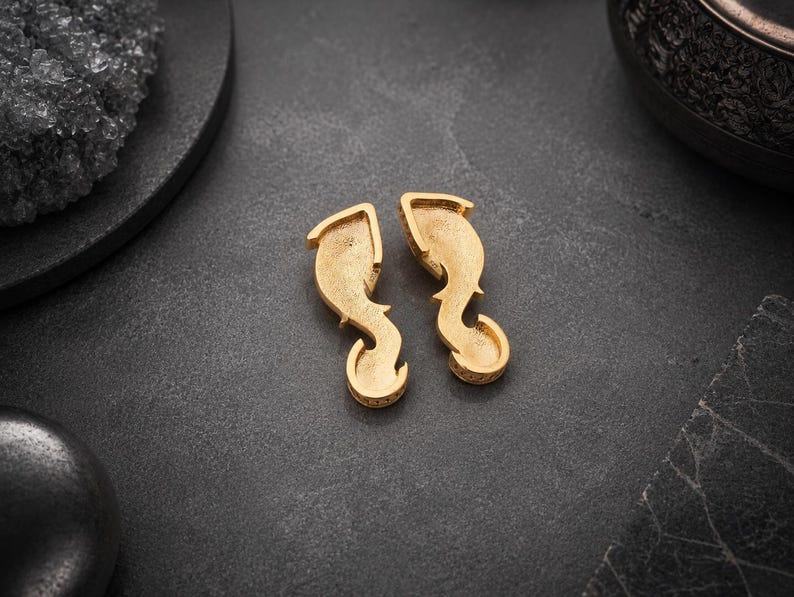 May include: A pair of gold-toned earrings featuring a curved, elongated shape with a textured, matte finish. The earrings have a hollow, recessed center design that tapers into a rounded, hook-like bottom section, presenting a distinct metallic luster.