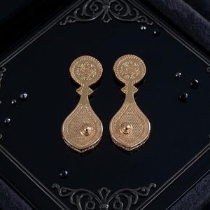May include: A pair of gold-toned drop earrings featuring a circular top section with an intricate embossed pattern and a larger, teardrop-shaped bottom section with a textured, mesh-like surface and a small central stud detail on each piece.