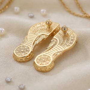 May include: A pair of gold-toned metal shoe-shaped ornaments featuring intricate embossed floral and geometric patterns, each topped with a small spherical stud. The items are surrounded by scattered white pearls and gold-toned chain necklaces.