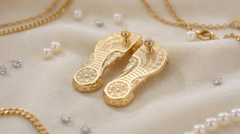 May include: A pair of gold-toned metal shoe-shaped ornaments featuring intricate embossed floral and geometric patterns, each topped with a small spherical stud. The items are surrounded by scattered white pearls and gold-toned chain necklaces.