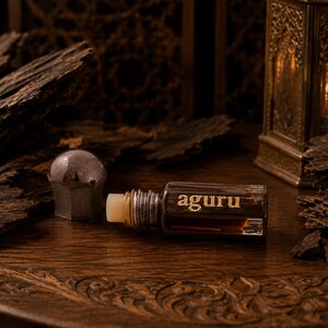 May include: A small rectangular glass bottle of dark brown liquid with a silver cap, featuring the word aguru printed in gold on the front. The bottle is positioned horizontally next to a detached silver cap and pieces of dark textured wood.
