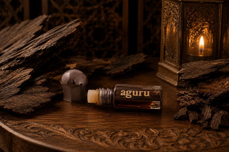 May include: A small rectangular glass bottle of dark brown liquid with a silver cap, featuring the word aguru printed in gold on the front. The bottle is positioned horizontally next to a detached silver cap and pieces of dark textured wood.