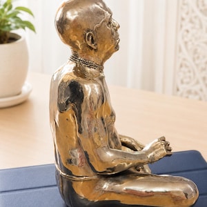 May include: A polished bronze-colored sculpture of a seated figure in a meditative pose. The figure is bald, has a prominent nose, and wears a beaded necklace. The sculpture has a smooth, reflective metallic finish and is shown from a side profile.