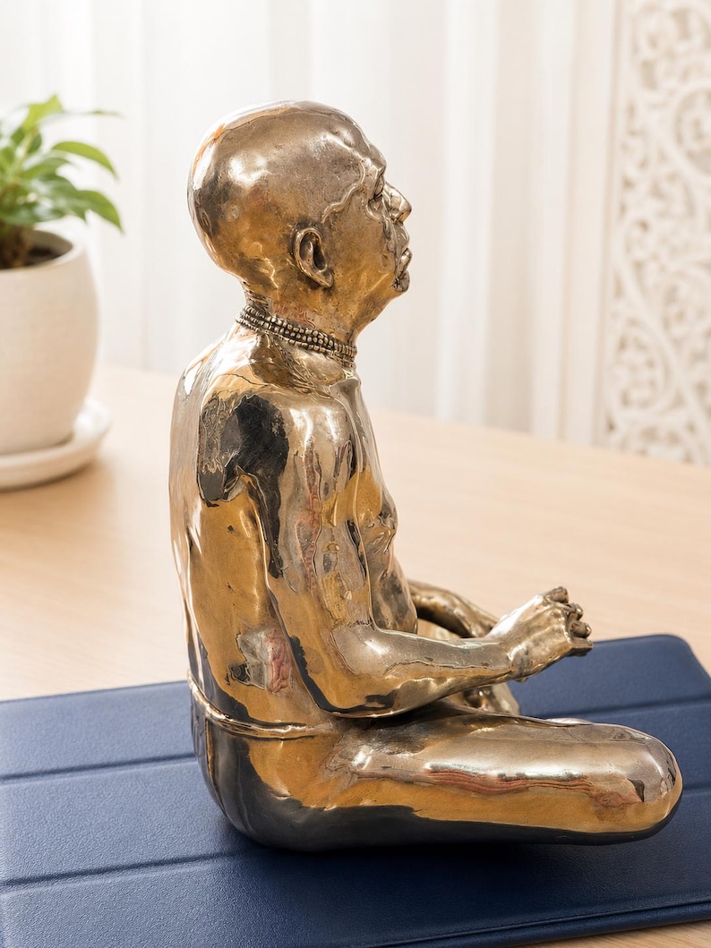 May include: A polished bronze-colored sculpture of a seated figure in a meditative pose. The figure is bald, has a prominent nose, and wears a beaded necklace. The sculpture has a smooth, reflective metallic finish and is shown from a side profile.