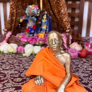 May include: A metallic bronze-toned statue of a seated figure with a shaved head, draped in a bright orange cloth. The figure has a serious expression and is positioned in front of a small, ornate wooden shrine containing two smaller colorful deities.