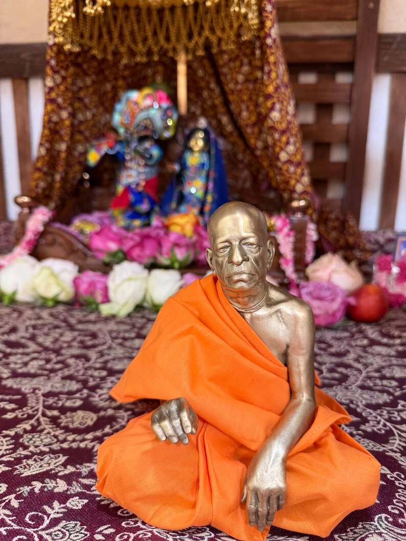 May include: A metallic bronze-toned statue of a seated figure with a shaved head, draped in a bright orange cloth. The figure has a serious expression and is positioned in front of a small, ornate wooden shrine containing two smaller colorful deities.