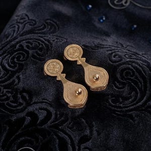 May include: A pair of gold-toned metal earrings featuring a circular top section with an embossed pattern and a teardrop-shaped lower section with a central raised sphere. The earrings have a textured, metallic finish and a distinct, elongated shape.