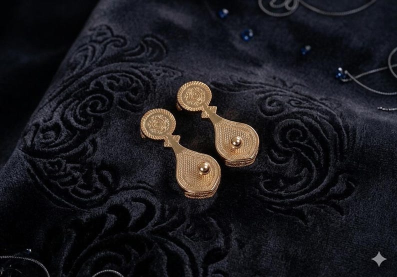 May include: A pair of gold-toned metal earrings featuring a circular top section with an embossed pattern and a teardrop-shaped lower section with a central raised sphere. The earrings have a textured, metallic finish and a distinct, elongated shape.