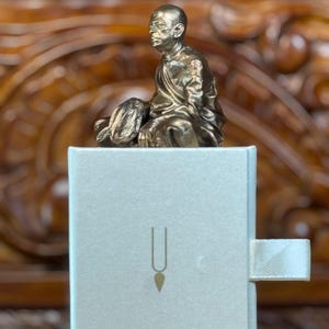 May include: A small, bronze-colored statue of a seated figure atop a light gray box. The box has a gold-colored symbol on the front and a white ribbon pull. The background is a carved wooden surface.