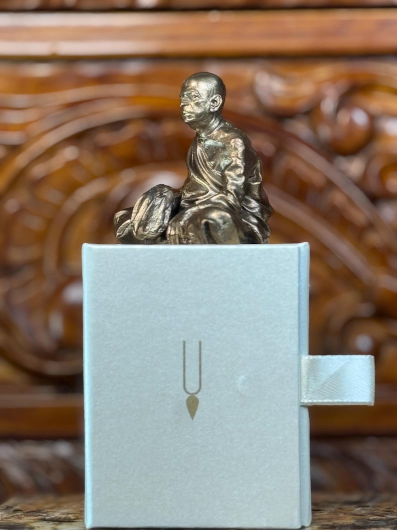 May include: A small, bronze-colored statue of a seated figure atop a light gray box. The box has a gold-colored symbol on the front and a white ribbon pull. The background is a carved wooden surface.