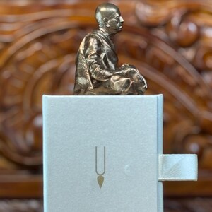 May include: A bronze-colored statue of a seated figure atop a white box with a gold-colored symbol. The statue is detailed, showing the person in a meditative pose. The box has a ribbon pull.