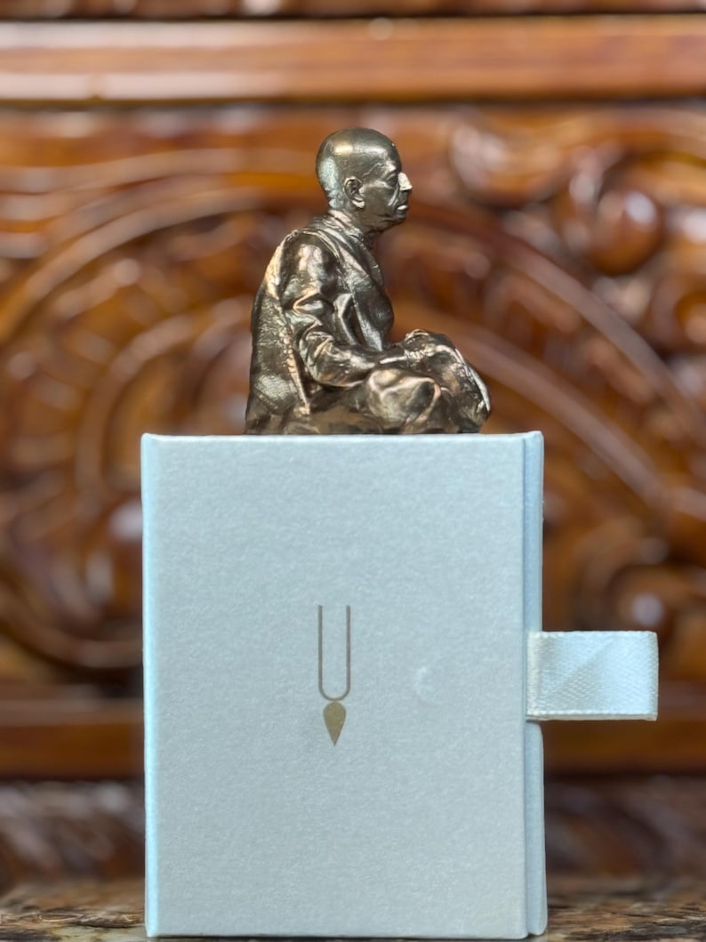 May include: A bronze-colored statue of a seated figure atop a white box with a gold-colored symbol. The statue is detailed, showing the person in a meditative pose. The box has a ribbon pull.