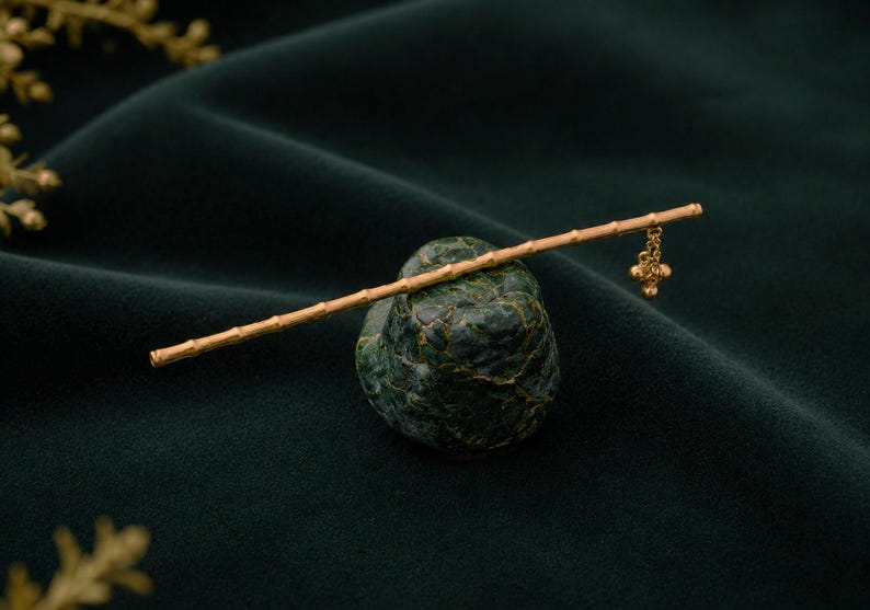Puede incluir: Un palillo para el cabello de tono dorado dise&ntilde;ado para parecerse a un tallo de bamb&uacute;, con una superficie texturizada con nudos segmentados. Una peque&ntilde;a y delicada cadena dorada con un dije colgante cuelga de un extremo, apoyado en una piedra verde.