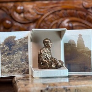 May include: A bronze-colored statue of a seated figure in a white box, with sepia-toned landscape photos on either side. The statue is of a person in a meditative pose. The background is a wooden surface.