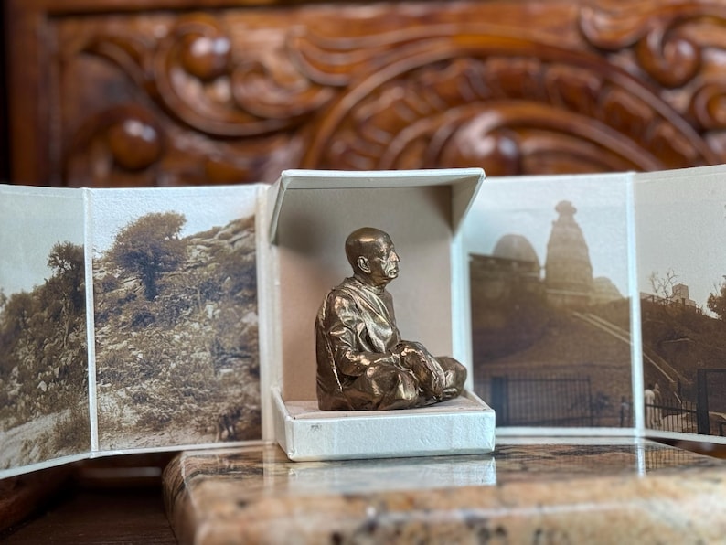 May include: A bronze-colored statue of a seated figure in a white box, with sepia-toned landscape photos on either side. The statue is of a person in a meditative pose. The background is a wooden surface.