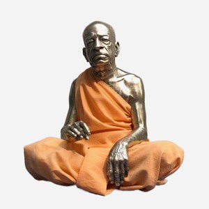 May include: A metallic silver-toned sculpture of a seated figure with a shaved head, depicted in a meditative pose. The figure is draped in a draped orange fabric garment that covers the torso and legs, leaving the arms and chest partially exposed.