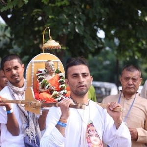May include: A small statue of a person in orange robes sits on a decorated wooden palanquin with a gold-colored canopy. The palanquin is supported by two wooden poles and is being carried by individuals in a procession outdoors.