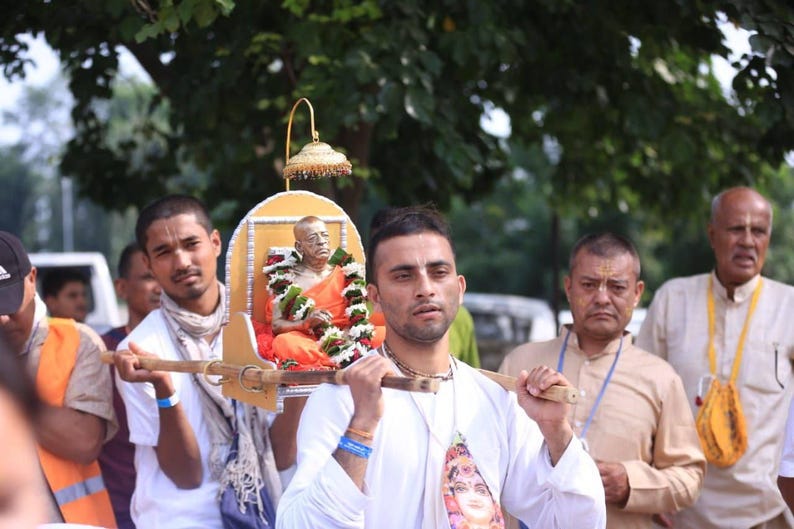 May include: A small statue of a person in orange robes sits on a decorated wooden palanquin with a gold-colored canopy. The palanquin is supported by two wooden poles and is being carried by individuals in a procession outdoors.