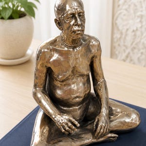 May include: A metallic bronze-toned sculpture of a seated person with a bald head and a mustache, depicted in a meditative pose. The figure has a textured surface, wears a simple beaded necklace, and is shown from the chest up in a seated position.