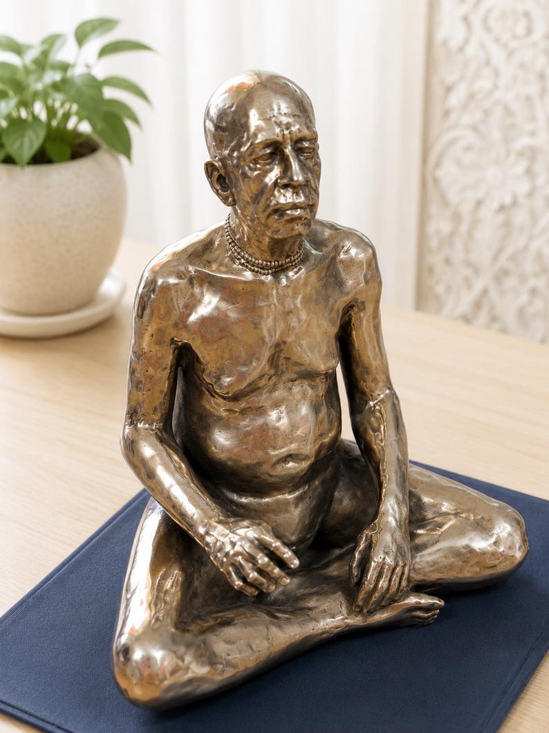May include: A metallic bronze-toned sculpture of a seated person with a bald head and a mustache, depicted in a meditative pose. The figure has a textured surface, wears a simple beaded necklace, and is shown from the chest up in a seated position.