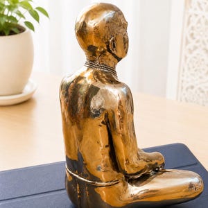 May include: A metallic gold-toned sculpture of a seated figure in a meditative pose. The figure has a smooth, reflective surface, a bald head, and a simple band around the neck. The sculpture is captured from a side-rear angle, showing the posture.