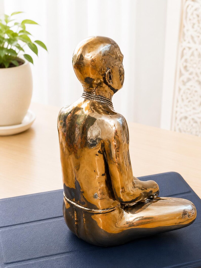 May include: A metallic gold-toned sculpture of a seated figure in a meditative pose. The figure has a smooth, reflective surface, a bald head, and a simple band around the neck. The sculpture is captured from a side-rear angle, showing the posture.