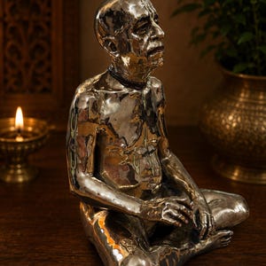 May include: A polished silver-toned figurine of a seated person with a shaved head, wearing a dark beaded necklace. The figure is depicted in a meditative pose with hands resting on the legs, featuring detailed facial expressions and markings.