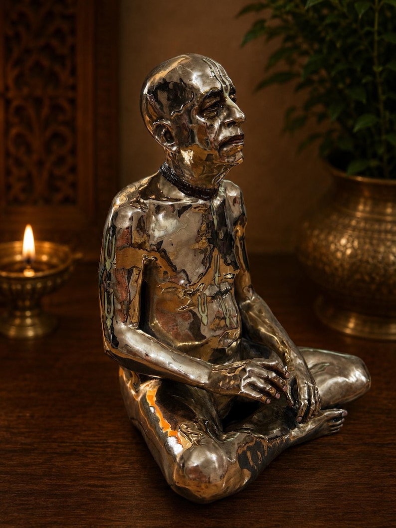 May include: A polished silver-toned figurine of a seated person with a shaved head, wearing a dark beaded necklace. The figure is depicted in a meditative pose with hands resting on the legs, featuring detailed facial expressions and markings.