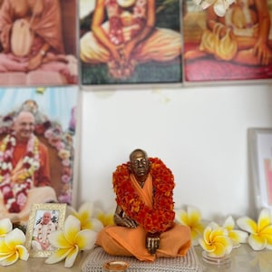 May include: A bronze statue of a seated figure wearing an orange robe and a garland of orange flowers. The statue is on a small, gray knitted mat, with small gold-colored cups and shoes in front. Framed portraits and white and yellow flowers are in the background.
