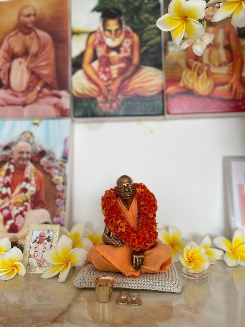 May include: A bronze statue of a seated figure wearing an orange robe and a garland of orange flowers. The statue is on a small, gray knitted mat, with small gold-colored cups and shoes in front. Framed portraits and white and yellow flowers are in the background.