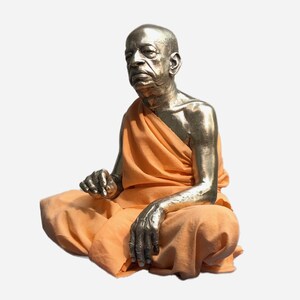 May include: A metallic, silver-toned sculpture of a seated figure with a shaved head, draped in a bright orange fabric garment. The figure is depicted in a cross-legged position with one hand resting on the knee and the other raised slightly.