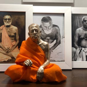 May include: A silver-colored statue of a seated figure in an orange robe, positioned in front of three framed black and white portraits. The statue is the focal point, with the portraits serving as a backdrop.