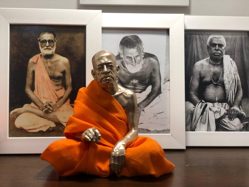 May include: A silver-colored statue of a seated figure in an orange robe, positioned in front of three framed black and white portraits. The statue is the focal point, with the portraits serving as a backdrop.