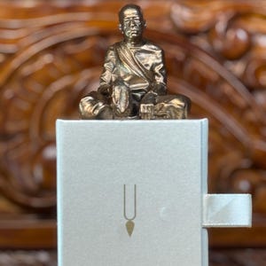 May include: A small, gold-colored statue of a seated figure atop a white box. The box has a simple design with a vertical line and a teardrop shape. The statue is detailed, and the box has a ribbon pull.