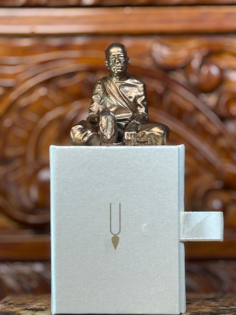 May include: A small, gold-colored statue of a seated figure atop a white box. The box has a simple design with a vertical line and a teardrop shape. The statue is detailed, and the box has a ribbon pull.