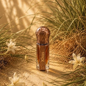 May include: A hexagonal glass perfume bottle with a dark amber liquid inside and a textured, metallic-looking cap. The word Khus is printed in white vertical lettering on the front face of the bottle, which has a clear glass base and sides.