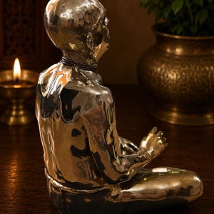 May include: A polished silver-toned metallic sculpture of a seated figure in a meditative pose. The figure has a smooth, reflective surface, a bald head, and a dark beaded necklace around the neck, captured from a side profile view.