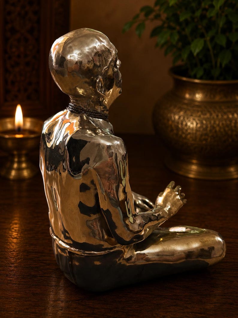 May include: A polished silver-toned metallic sculpture of a seated figure in a meditative pose. The figure has a smooth, reflective surface, a bald head, and a dark beaded necklace around the neck, captured from a side profile view.