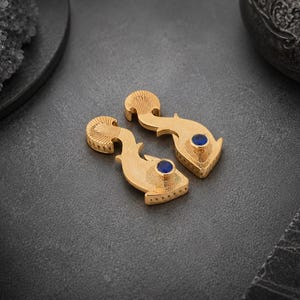 May include: A pair of gold-toned metal earrings shaped like stylized fish, featuring intricate etched line detailing on the fins and body, with a single round, deep blue stone set as the eye on each piece.