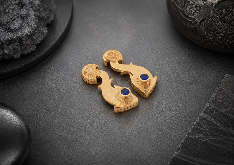 May include: A pair of gold-toned metal earrings shaped like stylized fish, featuring intricate etched line detailing on the fins and body, with a single round, deep blue stone set as the eye on each piece.