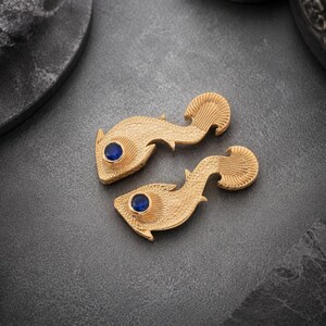 May include: A pair of gold-toned fish-shaped pins or brooches with a textured, scale-like surface finish. Each piece features a single, round, deep blue gemstone set as an eye. The design includes detailed fin markings and a curved tail section.