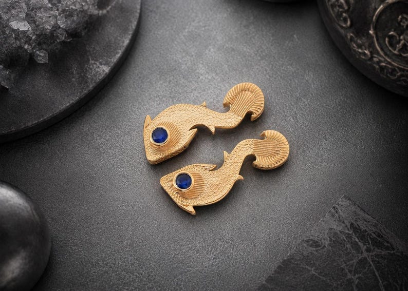 May include: A pair of gold-toned fish-shaped pins or brooches with a textured, scale-like surface finish. Each piece features a single, round, deep blue gemstone set as an eye. The design includes detailed fin markings and a curved tail section.