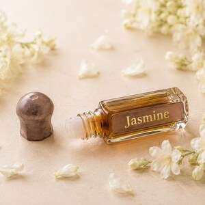 May include: A rectangular glass roller bottle filled with amber-colored liquid, featuring a metallic cap and the word Jasmine printed on the front. The bottle is positioned horizontally next to its removed cap, surrounded by small white flowers.
