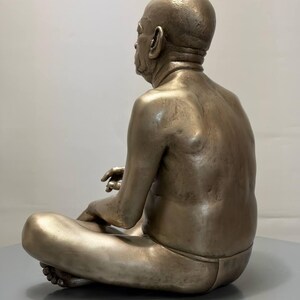 May include: A metallic, bronze-toned sculpture of a seated figure with a shaved head, depicted in a cross-legged position. The figure has a smooth, muscular back and is wearing simple, form-fitting lower-body garment, captured from a side profile.