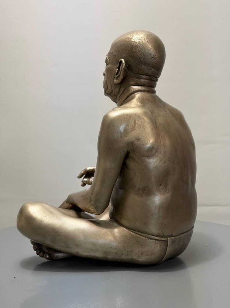 May include: A metallic, bronze-toned sculpture of a seated figure with a shaved head, depicted in a cross-legged position. The figure has a smooth, muscular back and is wearing simple, form-fitting lower-body garment, captured from a side profile.