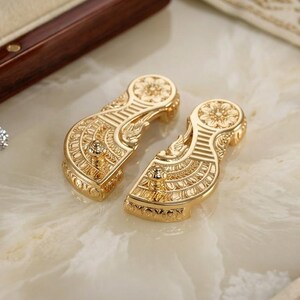 May include: Two gold-toned metal components featuring intricate engraved patterns, including floral motifs and linear textures. Each piece has a rounded top and a tapered base. Several small, loose faceted gemstones in blue and clear white are scattered nearby.