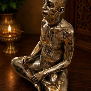 May include: A metallic silver-toned sculpture of a seated figure in a meditative pose. The figure has a shaved head, a beaded necklace, and visible markings on the forehead and chest. The surface of the sculpture has a hammered, reflective finish.