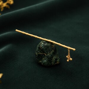 Puede incluir: Un palillo para el cabello de tono dorado con un dise&ntilde;o texturizado similar al bamb&uacute;. El accesorio incluye una peque&ntilde;a cadena colgante con tres diminutos dijes esf&eacute;ricos en un extremo, apoyado horizontalmente sobre una base de piedra verde.