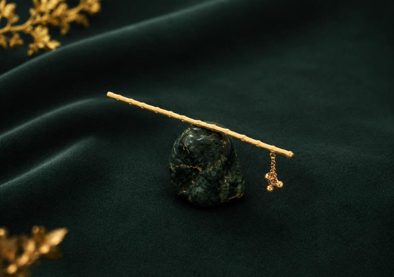 Puede incluir: Un palillo para el cabello de tono dorado con un dise&ntilde;o texturizado similar al bamb&uacute;. El accesorio incluye una peque&ntilde;a cadena colgante con tres diminutos dijes esf&eacute;ricos en un extremo, apoyado horizontalmente sobre una base de piedra verde.
